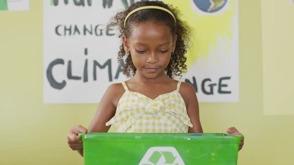 Video of happy african american girl holding box with recycling symbol in classroom