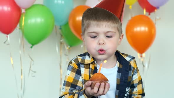 Boy in Party Cone Blows Candle on Cupcake Against Balloons