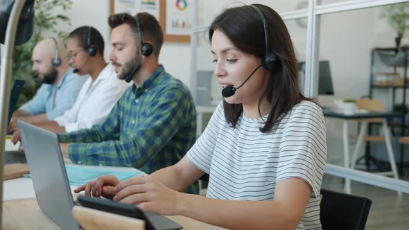 Cheerful Young Woman Call Center Worker Talking and Typing Using Modern Equipment with Colleagues in