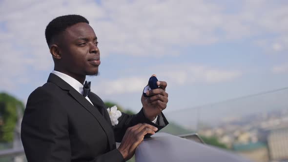 Side View of Thoughtful African American Groom in Wedding Suit Standing on Bridge Holding Wedding