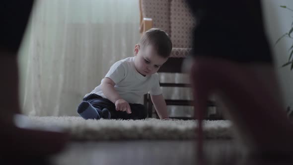 Cute Baby Boy Sitting on the Floor on Fluffy Carpet Playing Alone While His Young Mother Walking in