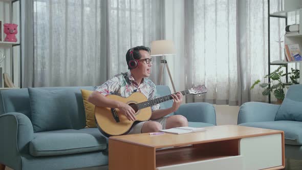 Asian Man Composer With Notebook On Table Wearing Headphones Singing And Playing Guitar At Home