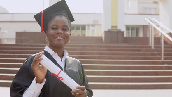 Young African American Female Graduate Standing in Front of the Camera with a Diploma and Books in