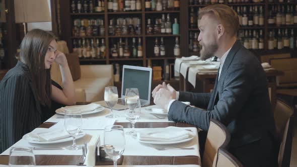 Young Beautiful Woman and Mature Man with Beard Having Business Dinner with Laptop Computer in