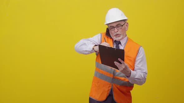 elderly Man engineer in construction uniform speaks on the phone and writes with pen on a paper tabl