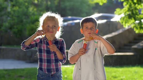 Two Little Children Boy and Girl Playing Outdoors and Blowing Soap Bubbles in Backlight Close Up