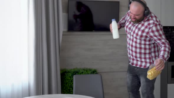 A Bearded Man in Wireless Headphones Walks Through the Kitchen with a Bottle of Milk and Dances