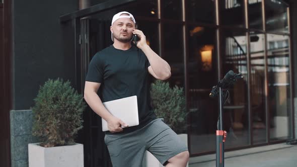 Full shot of busy man with e-scooter parked nearby talking on phone while working on laptop sitting