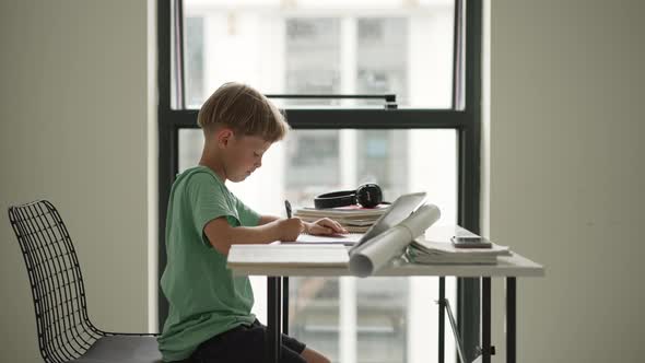 Caucasian Boy Sitting at Table with Workbook and Looking at Tablet Screen While Attentively