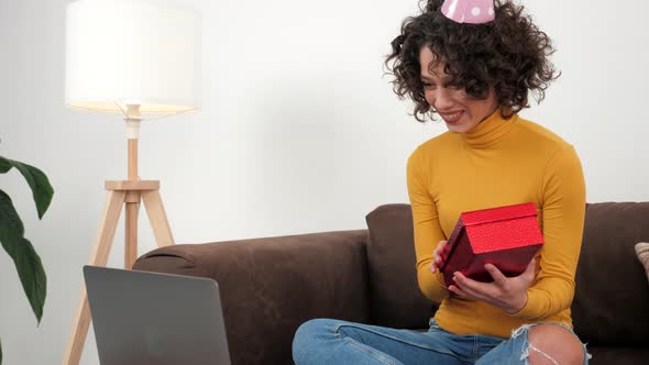 Happy Woman in Party Hat Opens Gift Box and Surprised Sitting in Couch at Home