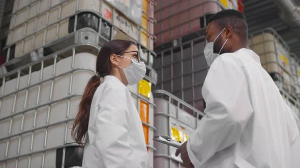 African and Caucasian Inspectors in Safety Mask and Lab Coat Standing in Chemical Factory Warehouse