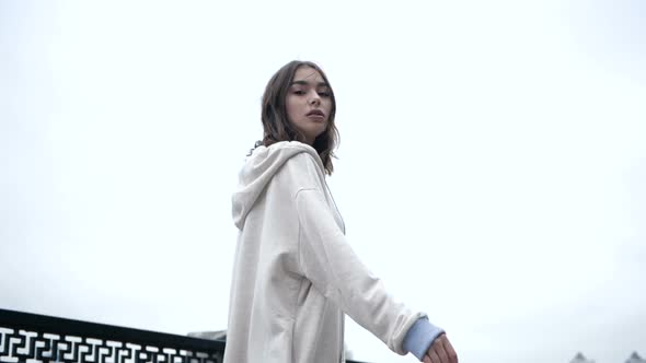 A Young Woman in a White Hoodie Walks Along the Embankment in the Early Morning