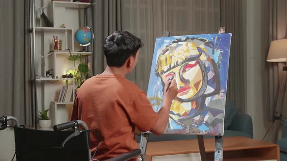 Hind View Of An Asian Artist Boy In Wheelchair Thinking And Painting A Girl's Face On The Canvas