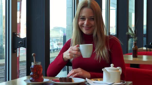 A Young Attractive Woman Sits at a Table with Meal in a Cafe, Drinks Tea and Smiles at the Camera