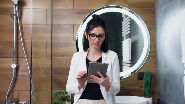 Brunette in Glasses Dressed in white Suit Working on Tablet PC Near the Bathroom's Mirror