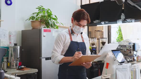 Asian cafe business owner wear protective mask, check order from screen on counter in restaurant.