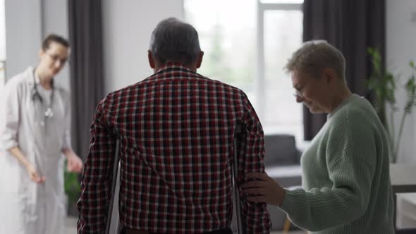 Old Man with Crutches Helped By His Wife and Doctor