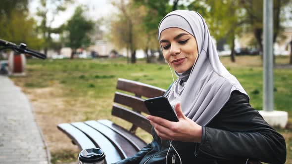 Portrait of Young Moderm Attractive Muslim Woman Sitting in Park Wearing Hijab and Listening to