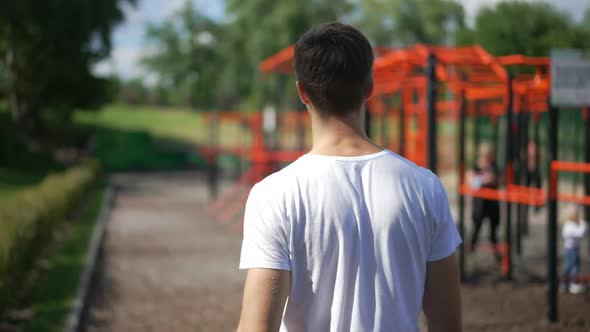 Back View Young Caucasian Man Walking in Slow Motion to Outdoors Exercise Equipment on Sunny Day
