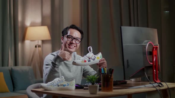 Smiling Asian Male Footwear Designer Holding Colourful Pattern Sneaker And Showing Thumbs Up Gesture