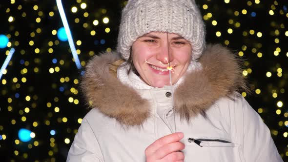 A Woman Holds a Sparkler and Smiles Standing Outside a Christmas Tree in the Open Air