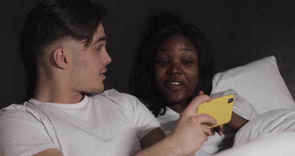 Happy Multi-ethnic Couple Watching Video Together on the Yellow Smartphone Lying in Bed at Night