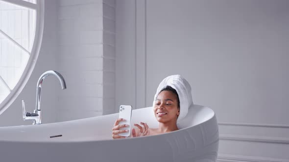 Black Woman Taking a Bath Looking at Phone with Her Hair Wrapped in a Towel