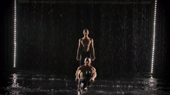 Two Young Athletes Demonstrate a Handstand. Male Acrobats with a Naked Torso Under Streams of Water