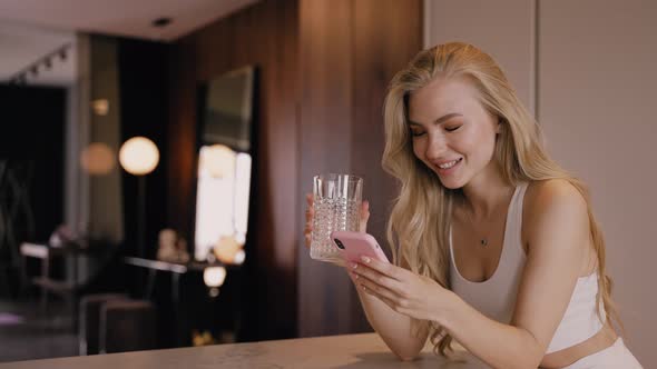 A Young Blonde in the Kitchen Drinks Water From a Glass and Chats By Phone