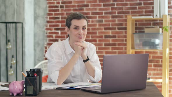 Nice Woman in a White Shirt Sits at an Office Table Propped Up Chin in Arms and Smiling Looks Into