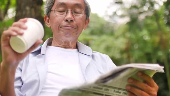 An old Asian man sits comfortably reading a book in a green park.