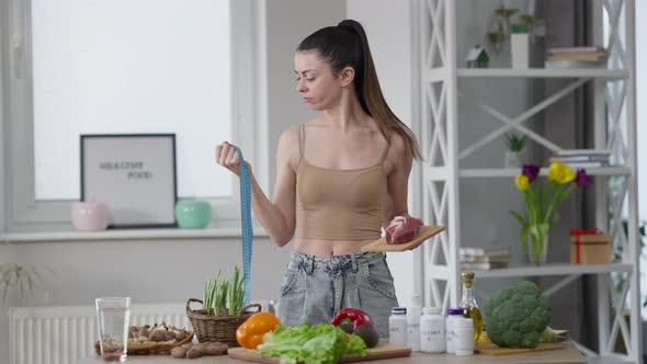 Unsure Caucasian Young Slim Woman Holding Measuring Tape and Raw Meat Making Decision and Looking at