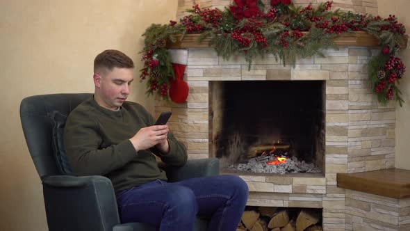A Young Man is Sitting in an Armchair By the Fireplace and Texting in a Smartphone