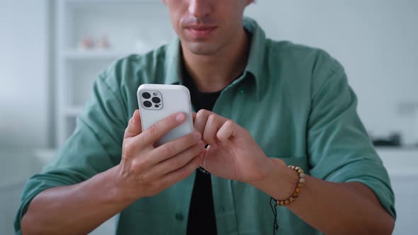 Smiling Young Man Sitting at a Table in the Kitchen and Using a Smartphone Scrolling Through the