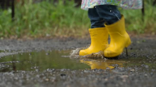 A child in yellow rubber boots runs and plays in a puddle of water in the rain