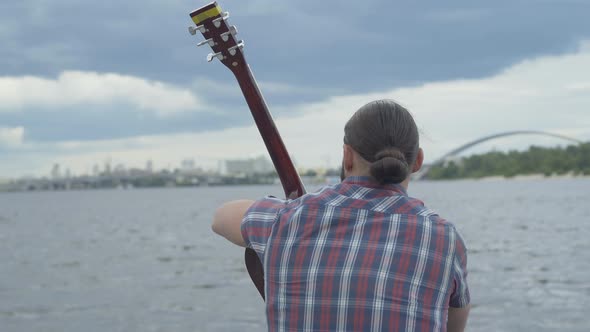 Back View of Caucasian Man Sitting with Guitar on River Bank with Blurred City at the Background