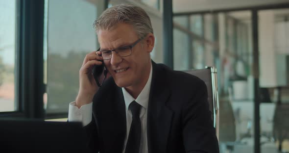 A businessman in formal suit and glasses talking on the phone, smiling, while working at the office.