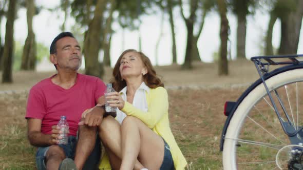 Couple Sitting on Lawn Enjoying Drinking Water After Cycling