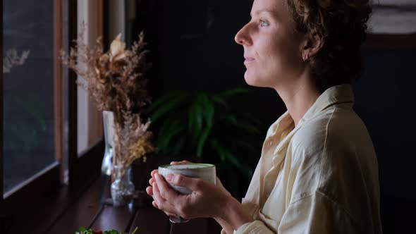 A Woman in Light Clothes Standing at the Window Looking Out the Window Breathing Air Holds a Cup