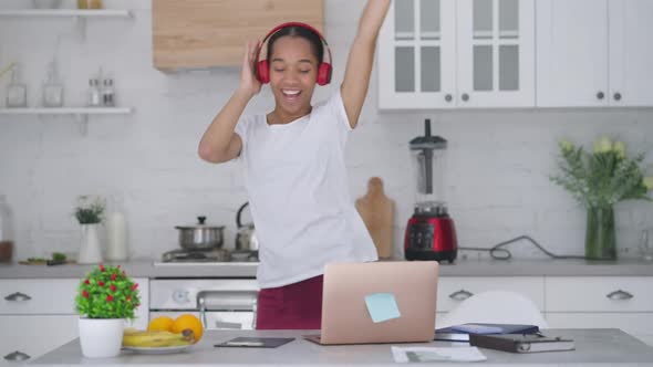 Joyful Satisfied Woman in Headphones Dancing in Kitchen Rejoicing Startup Profitability