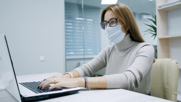 Woman Working at a Laptop in Protective Mask Typing Text in the Office Employee at Work on the