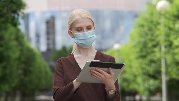 Business Woman in a Medical Mask Scrolls a Tablet in the City Center