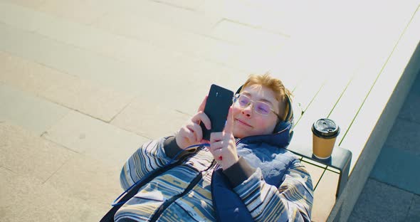 A Teenage Boy in Headphones During Online Chat While Lies on a Bench Whith Smartphone