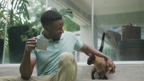 Happy african american man sitting with cat and drinking coffee in garden