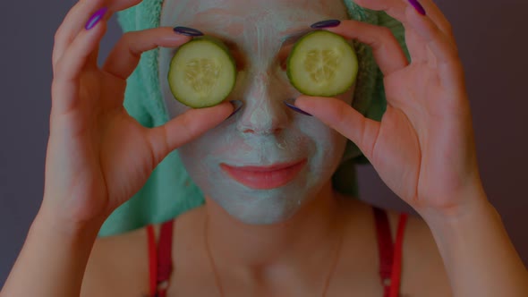 Young Woman with Towel on Head and Mask on Face Hiding Her Eyes Behind Cucumbers and Showing Them