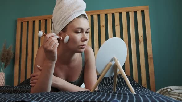 Young Woman Using Jade Facial Roller for Face Massage Lying on Bed in Bedroom Looking in the Mirror