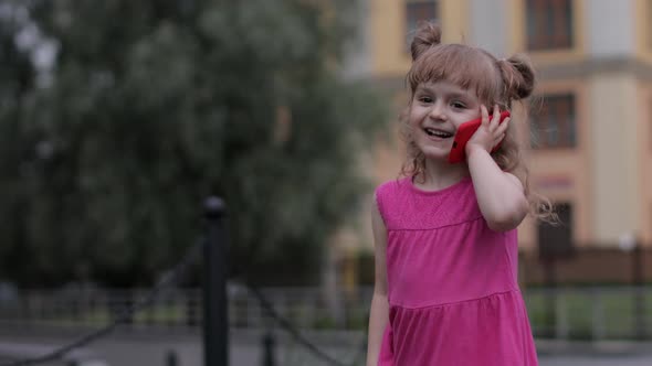 Child Girl Talking By Smartphone Outdoors. Kid in Pink Dress Talking By Mobile Phone on City Street