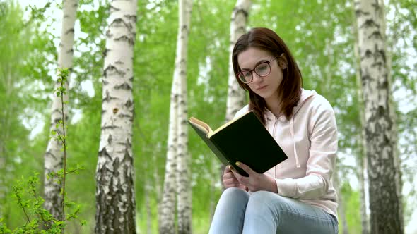 A Young Woman Is Sitting in Nature with a Book in Her Hands. A Girl Sits on a Stump in a Birch