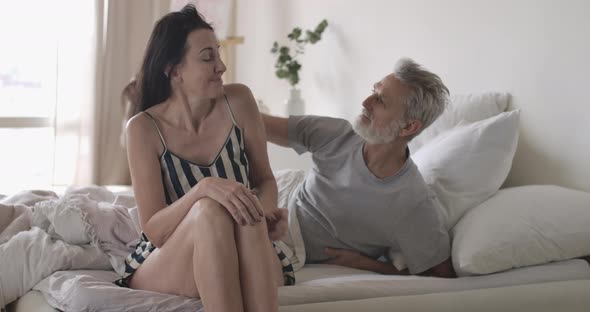 Handsome Smiling Caucasian Man Touching Wife's Hair in the Morning in Bed. Pleased Mature Woman