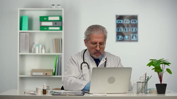 Aged Male Doctor in White Medical Coat Consults Remote Patient Online on a Laptop Computer